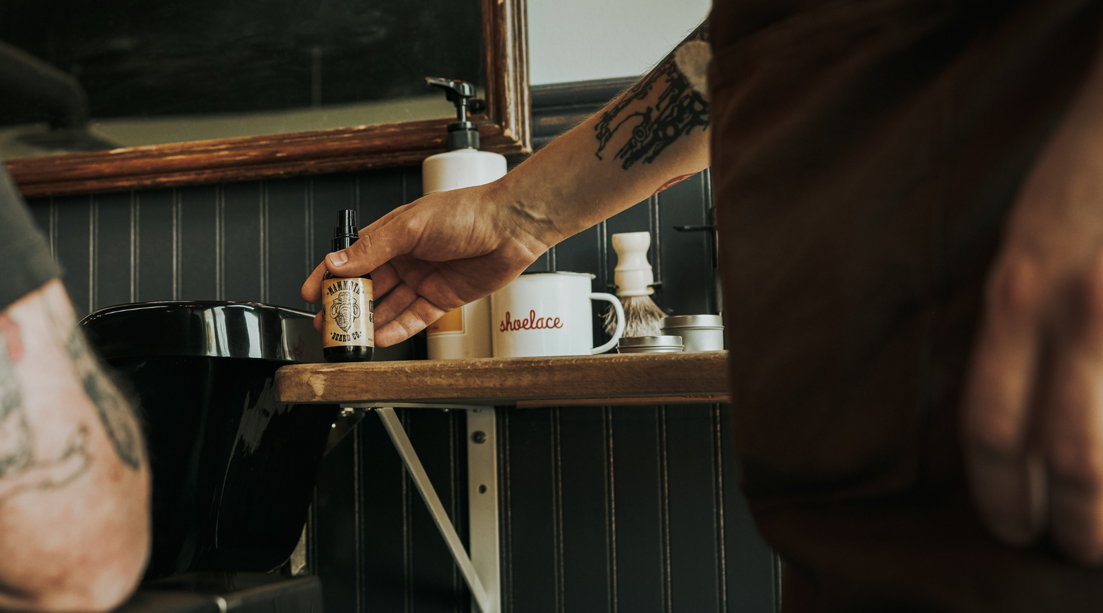 A tattooed barber’s hand reaching for a bottle of Mammoth Beard Co. beard oil.