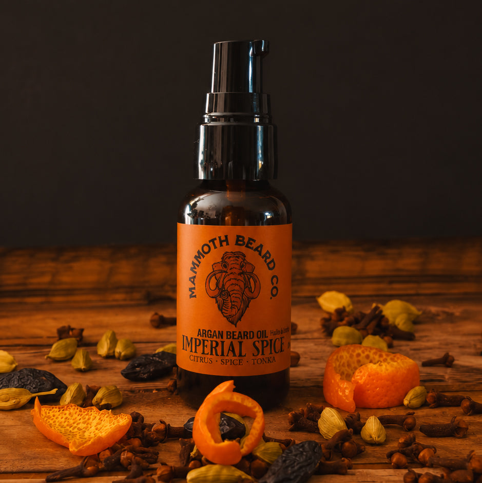 A bottle of Mammoth Beard Co. Argan Beard Oil in the Imperial Spice scent, styled on a rustic wooden surface surrounded by dried spices, cloves, and citrus peels against a dark background.