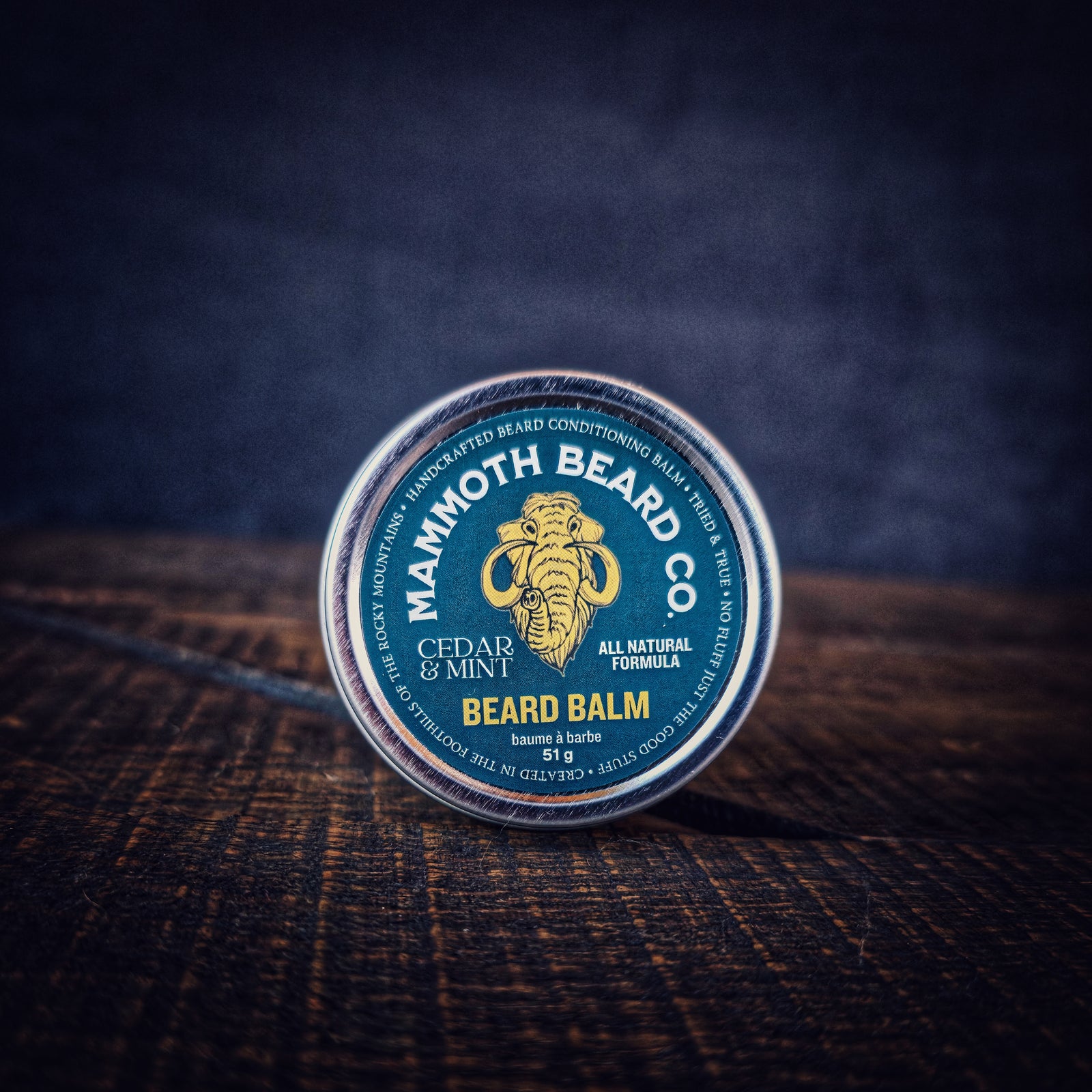 Closed tin of Cedar & Mint Beard Balm by Mammoth Beard Co on dark wood surface, natural beard conditioner with woods, mint and vanilla scent.
