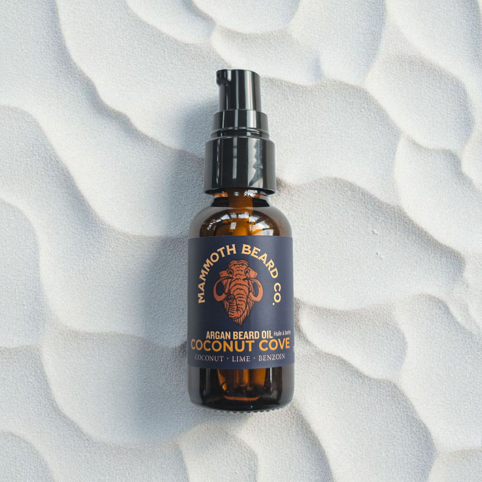 Mammoth Beard Co. Coconut Cove Argan Beard Oil bottle resting on a soft, rippled sand background, evoking a clean beach aesthetic and highlighting its tropical scent of coconut, lime, and benzoin.