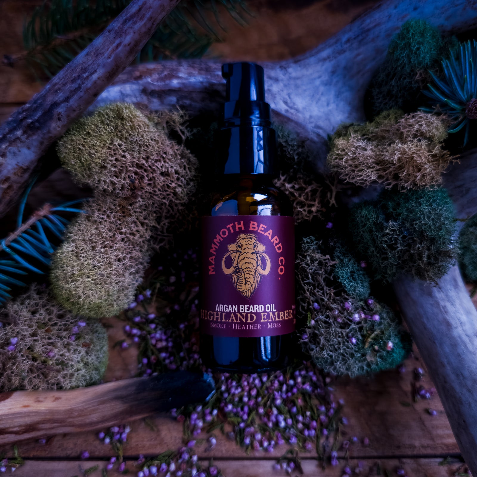 Amber bottle of Mammoth Beard Co. Highland Ember argan beard oil styled with moss, pine, antler, and scattered heather buds on weathered wood.