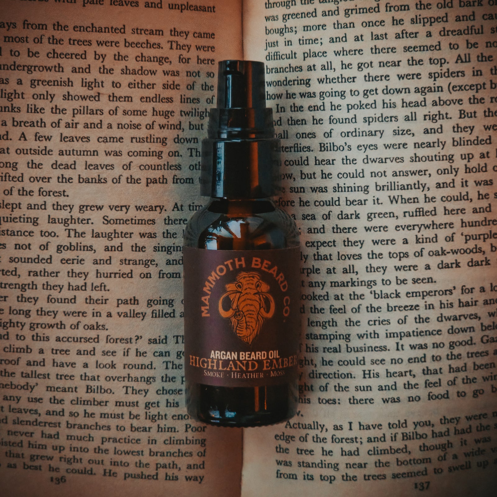 Mammoth Beard Co. Highland Ember argan beard oil in an amber glass pump bottle, standing on open book pages—moody, literary product shot.