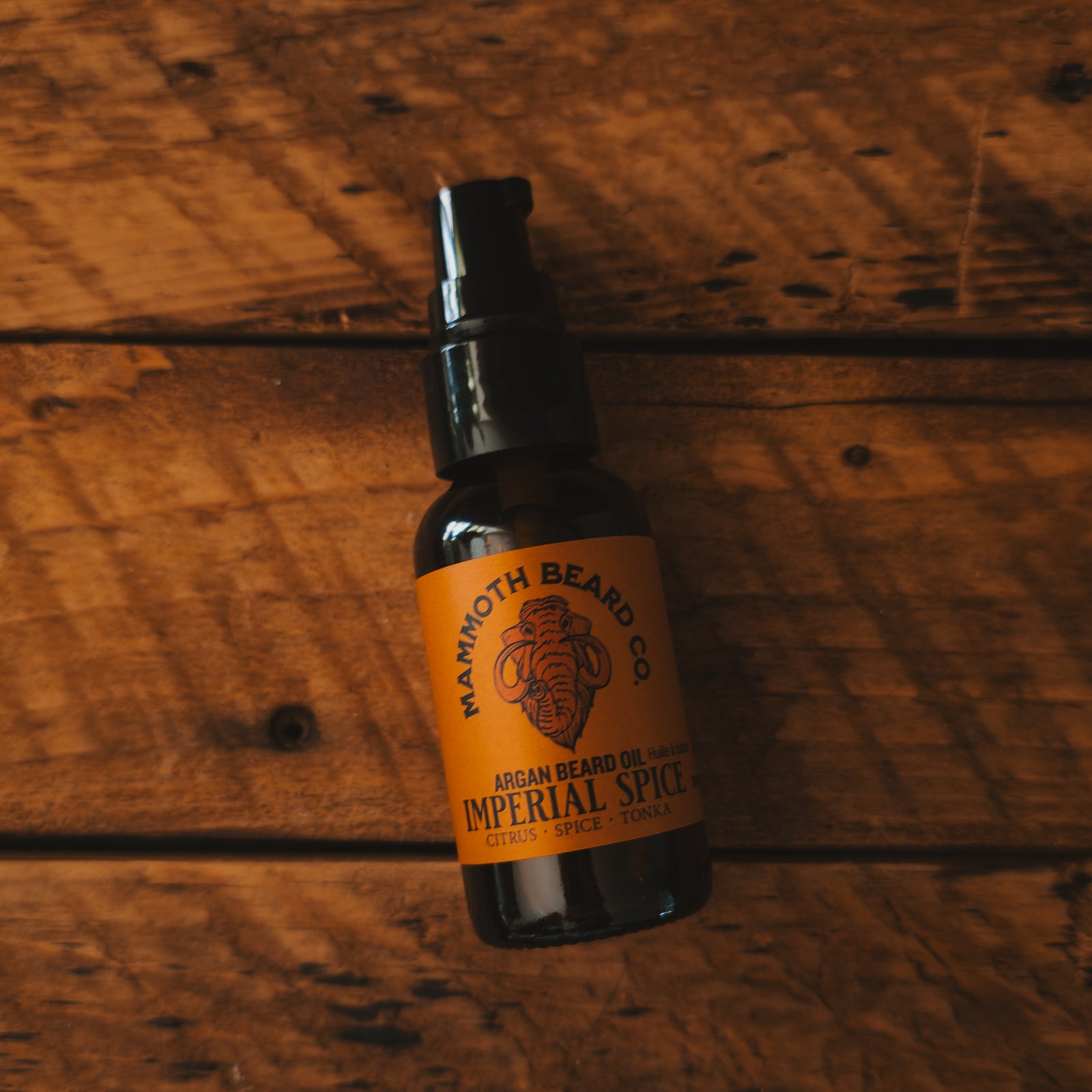 An amber bottle with a black pump cap and an orange label reading 'Mammoth Beard Co. Imperial Spice' lies on a rustic wooden surface.