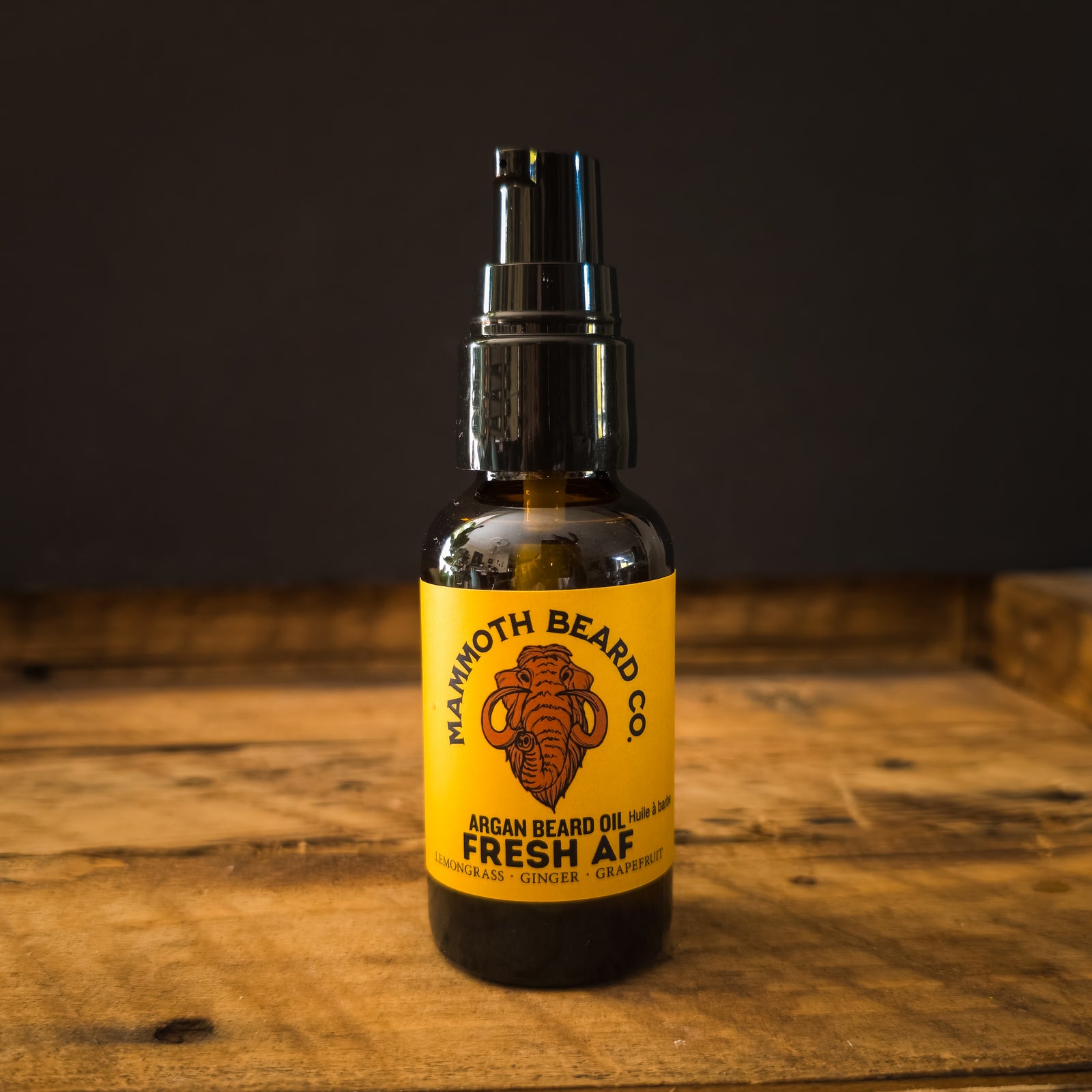 A bottle of Mammoth Beard Co. Fresh AF beard oil, featuring a black spray pump and a bright yellow label set on a rustic wooden surface against a dark background.