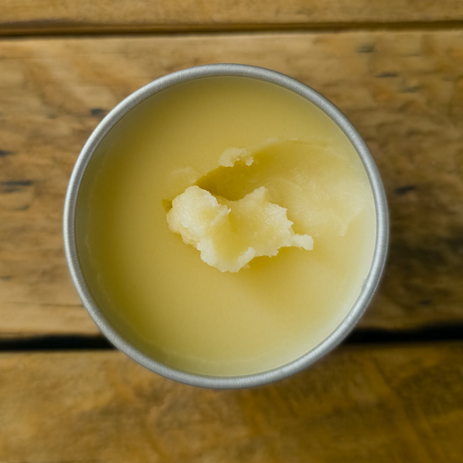 Top view of an open tin of Mammoth Beard Co. beard balm on a rustic wooden surface, highlighting its smooth, natural formula with a small portion scooped out.