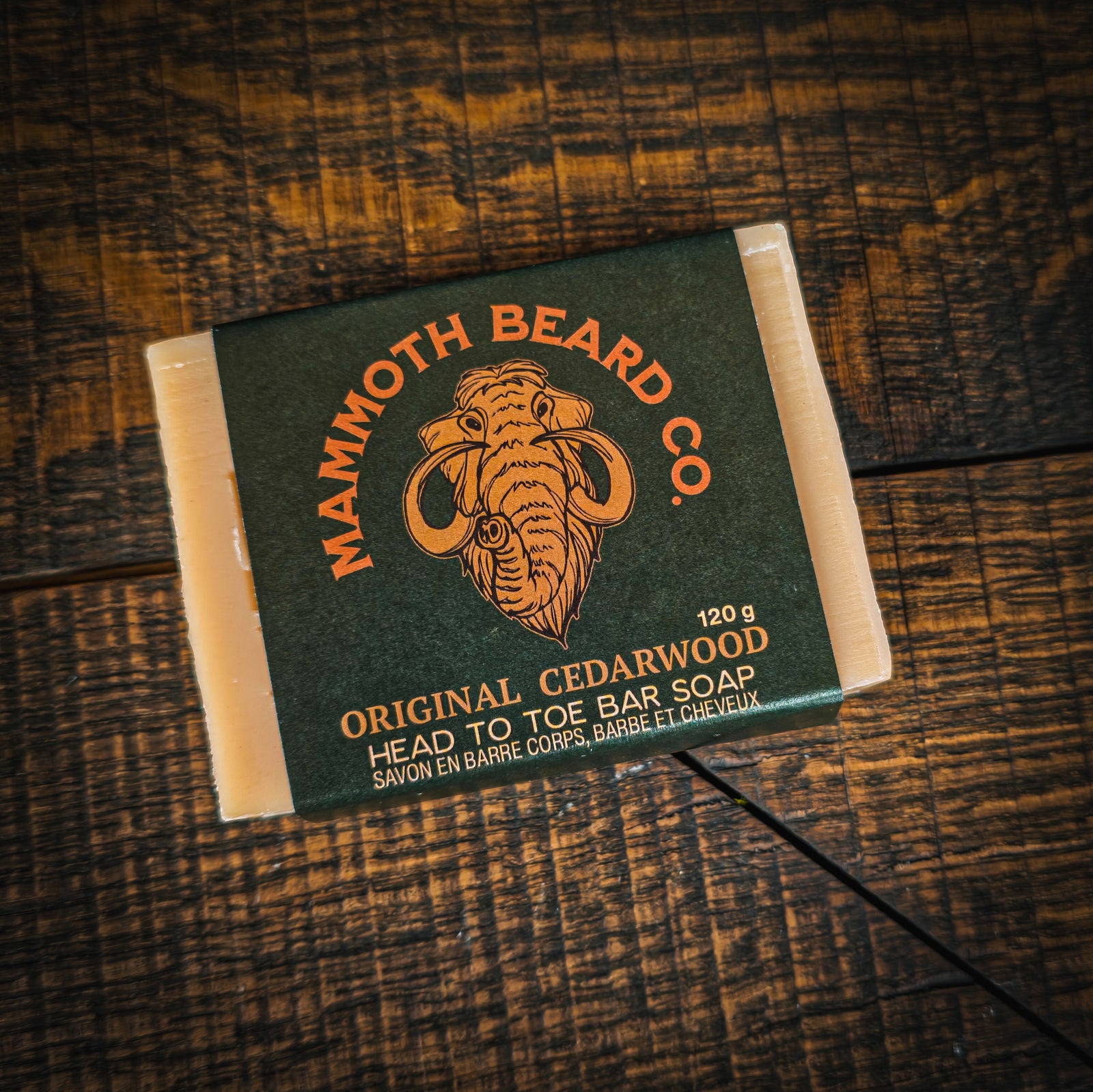 A rectangular bar of soap wrapped in a dark olive-green paper sleeve with orange text reading 'MAMMOTH BEARD CO. ORIGINAL CEDARWOOD HEAD TO TOE BAR SOAP' and an illustrated mammoth, placed on a dark wood surface.