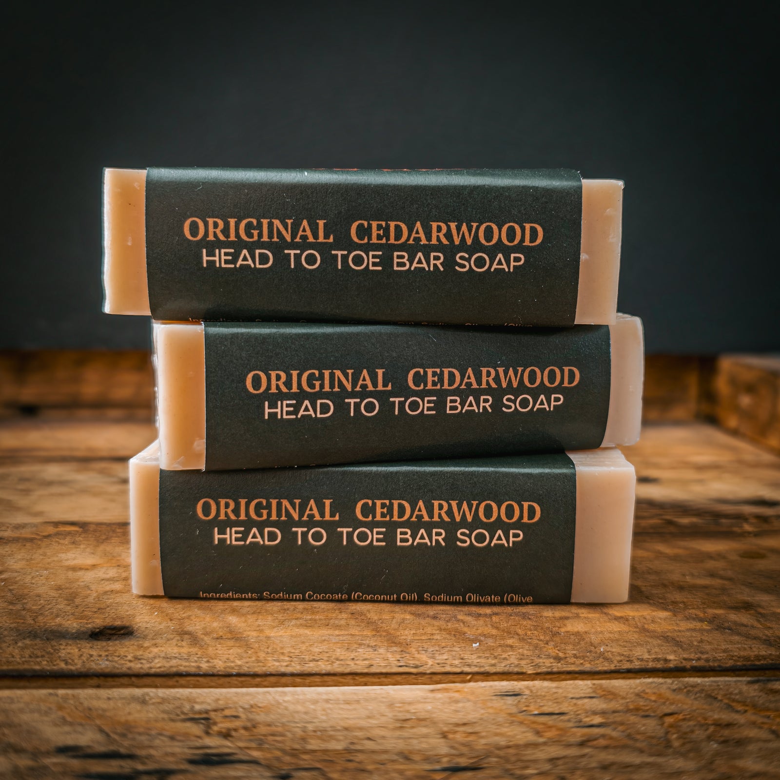 Three stacked bar soaps labeled 'Original Cedarwood Head to Toe Bar Soap' in beige with dark green paper bands, displayed on a rustic wooden surface against a dark background.