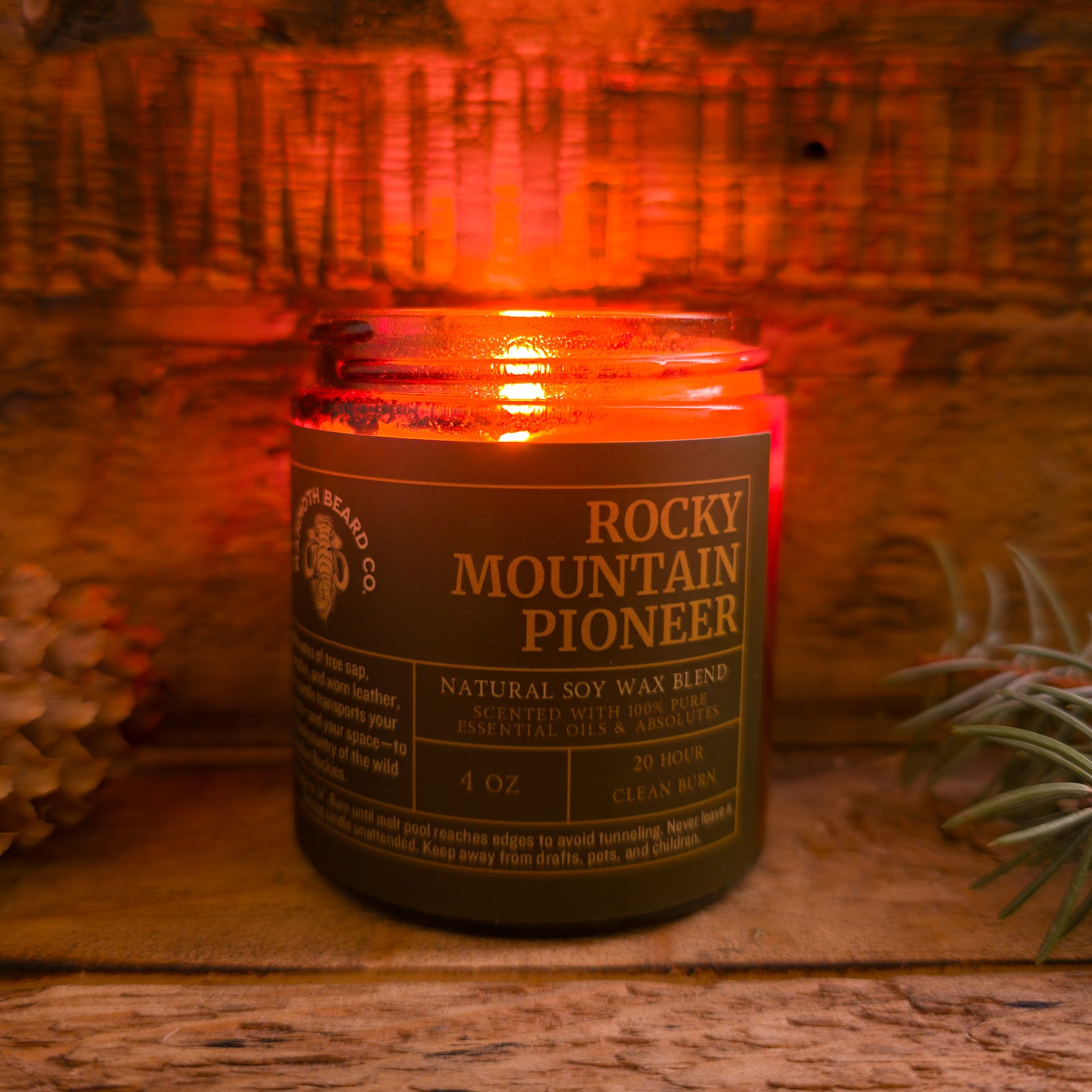 Rocky Mountain Pioneer candle burning, amber jar glowing warmly against a rustic wooden backdrop with pinecones and evergreen sprigs.