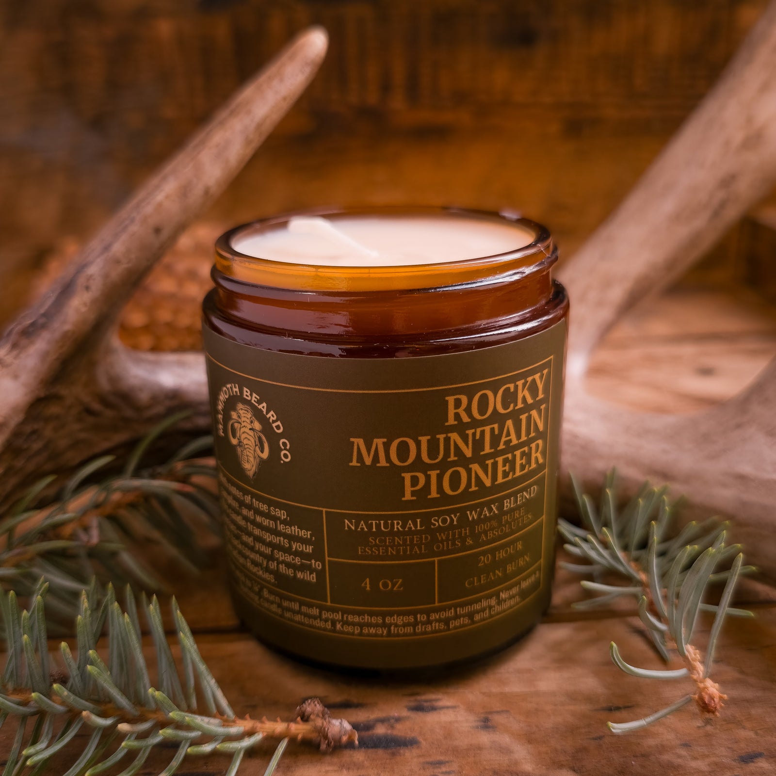 4 oz Rocky Mountain Pioneer soy wax candle with lid off, revealing creamy wax, displayed with antlers and pine greenery on a wooden surface.