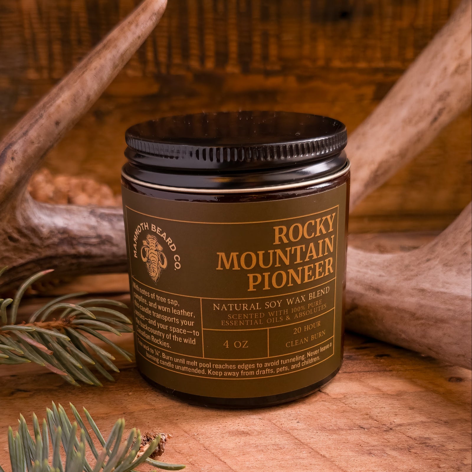 4 oz Rocky Mountain Pioneer natural soy wax candle with lid on, set against rustic wood and antlers, surrounded by evergreen sprigs.