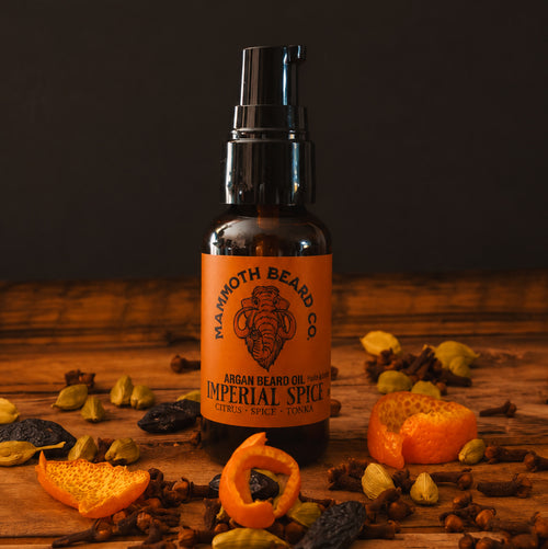 A bottle of Mammoth Beard Co. Argan Beard Oil in the Imperial Spice scent, styled on a rustic wooden surface surrounded by dried spices, cloves, and citrus peels against a dark background.