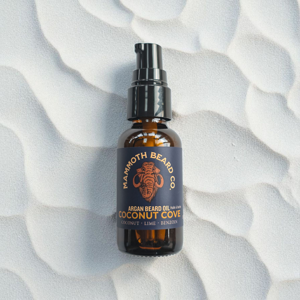 Mammoth Beard Co. Coconut Cove Argan Beard Oil bottle resting on a soft, rippled sand background, evoking a clean beach aesthetic and highlighting its tropical scent of coconut, lime, and benzoin.