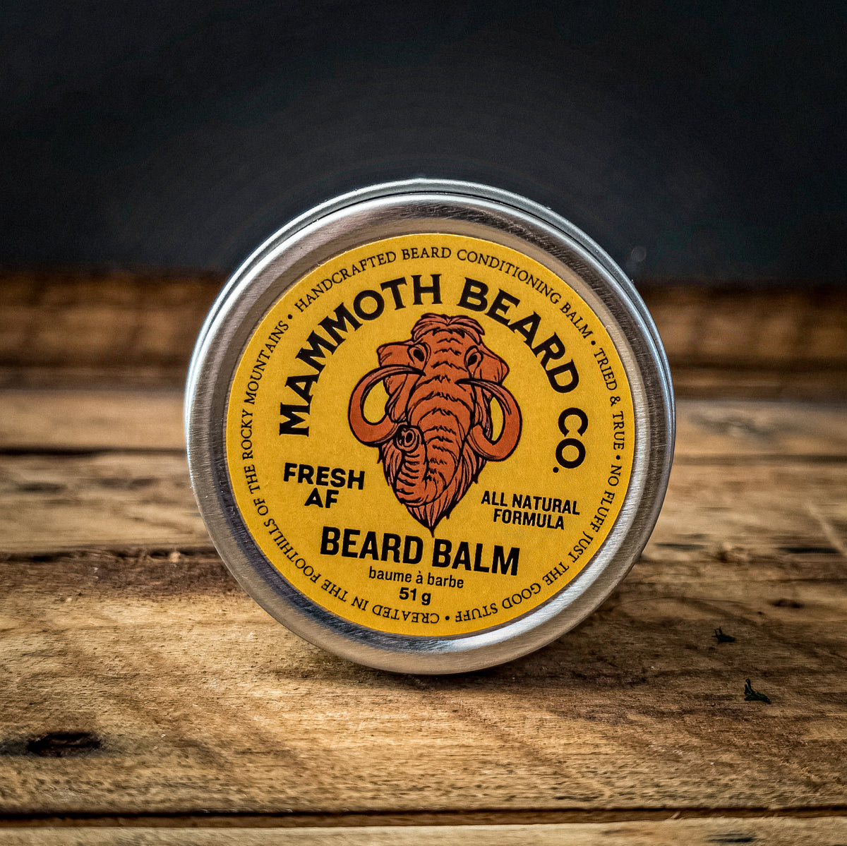 A round tin of Mammoth Beard Co. Fresh AF Beard Balm sits on a rustic wooden surface with a dark, moody background.