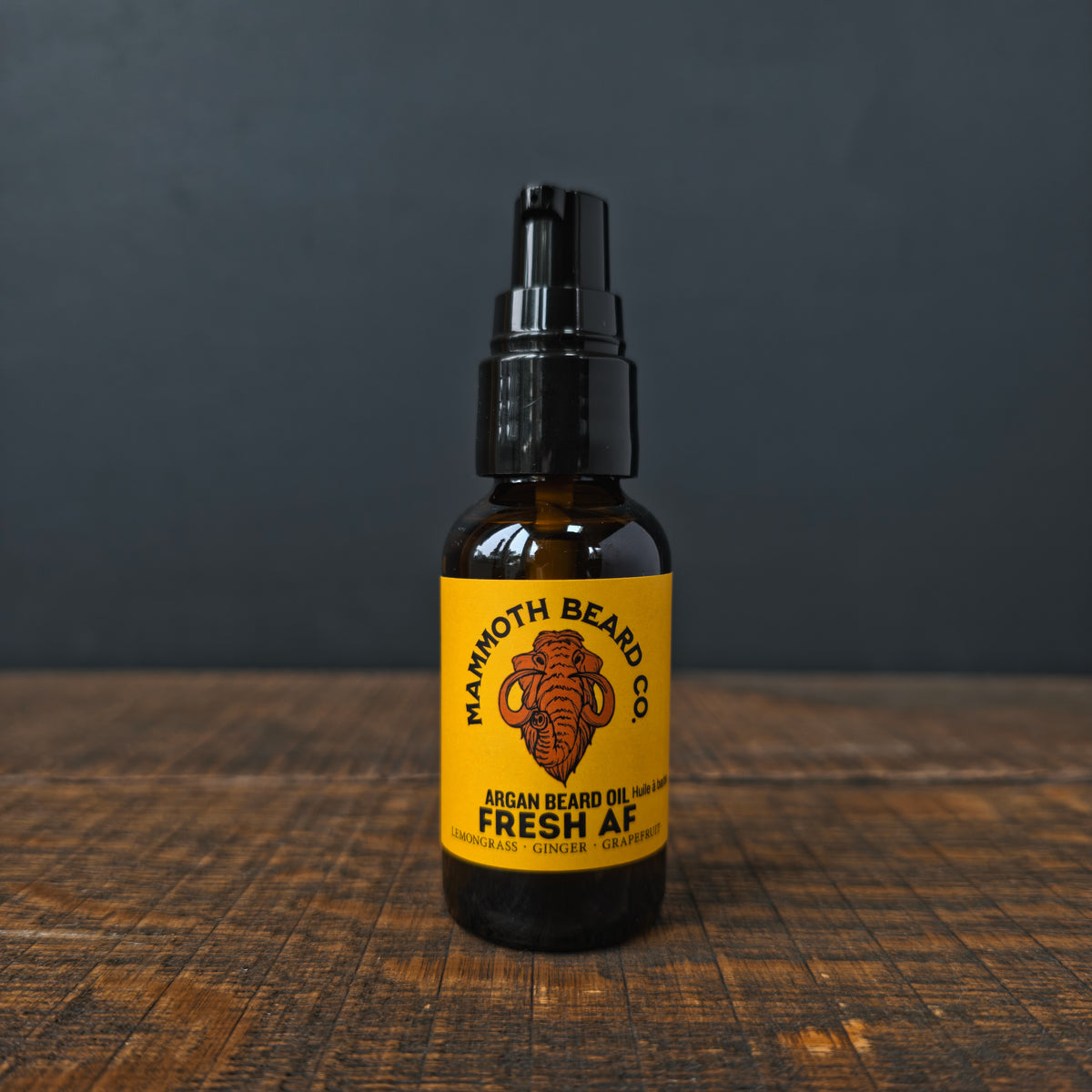 A bottle of Mammoth Beard Co. Argan Beard Oil in the 'Fresh AF' scent, featuring a black pump top and a bright yellow label set on a rustic wooden surface against a dark gray background.