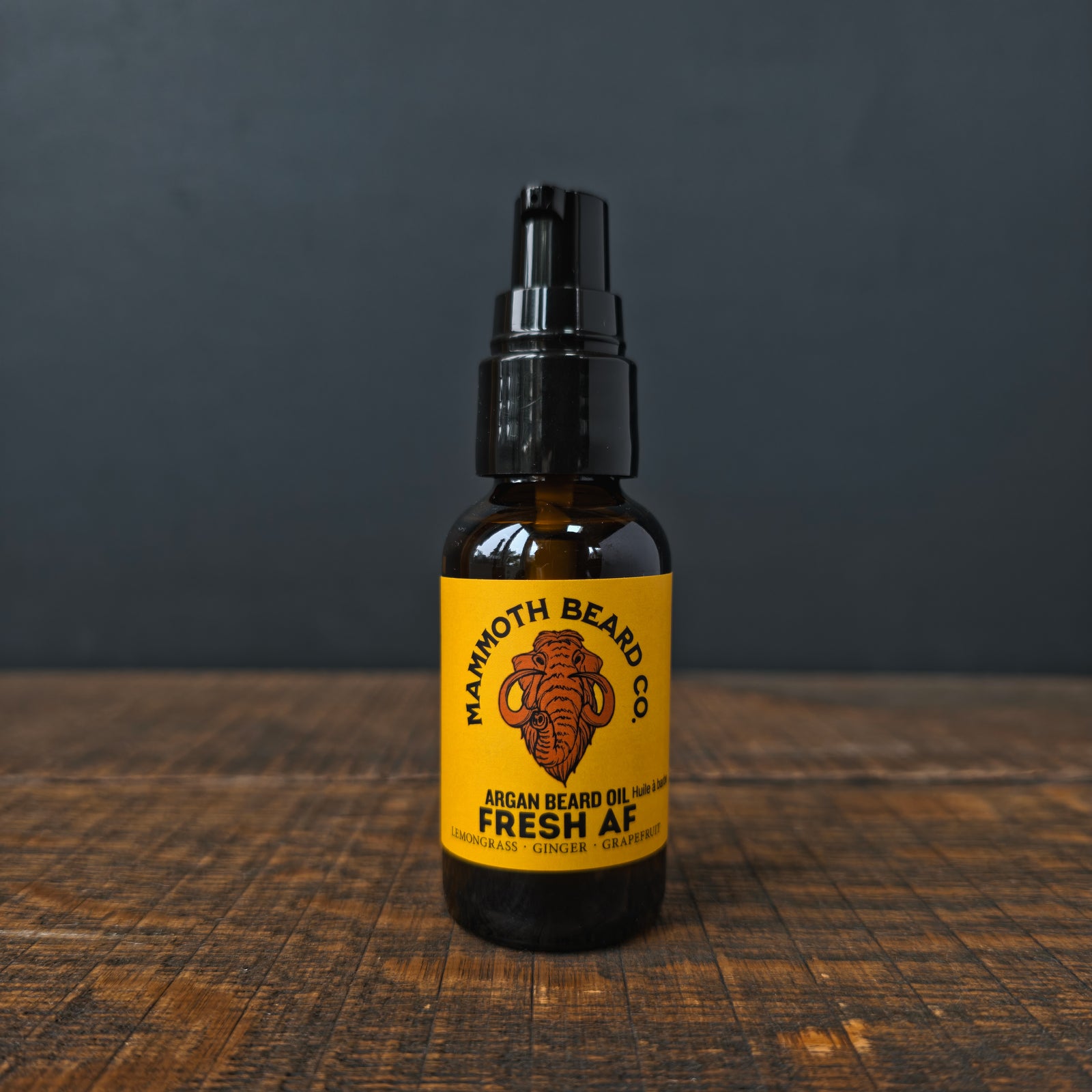 A bottle of Mammoth Beard Co. Argan Beard Oil in the 'Fresh AF' scent, featuring a black pump top and a bright yellow label set on a rustic wooden surface against a dark gray background.