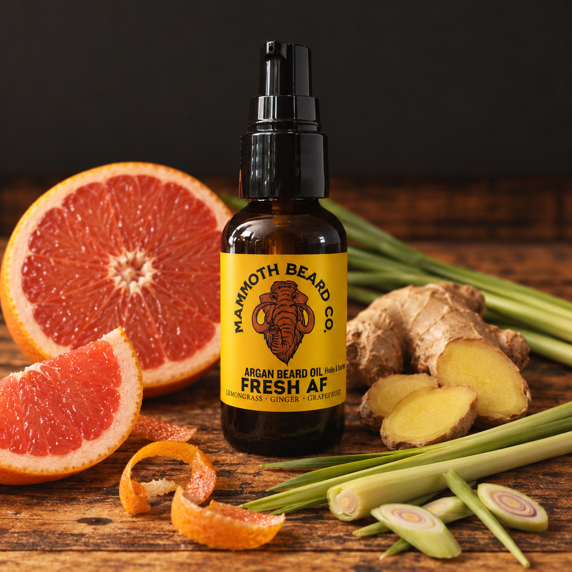 A bottle of Mammoth Beard Co. Fresh AF Beard Oil in an amber glass bottle with a black pump top and a yellow label, styled on a rustic wooden surface with sliced grapefruit, lemongrass, and ginger roots.