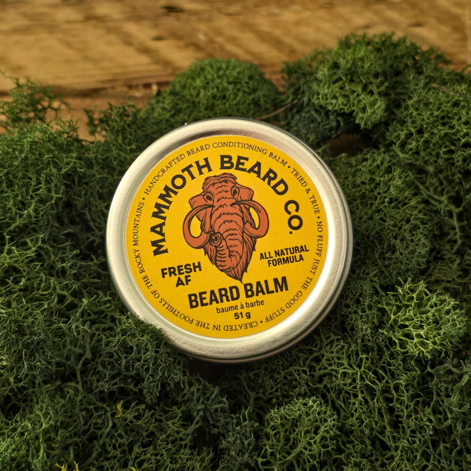 A round metal tin of Mammoth Beard Co. Fresh AF Beard Balm sits on a bed of green moss, with a rustic wooden surface visible in the background.