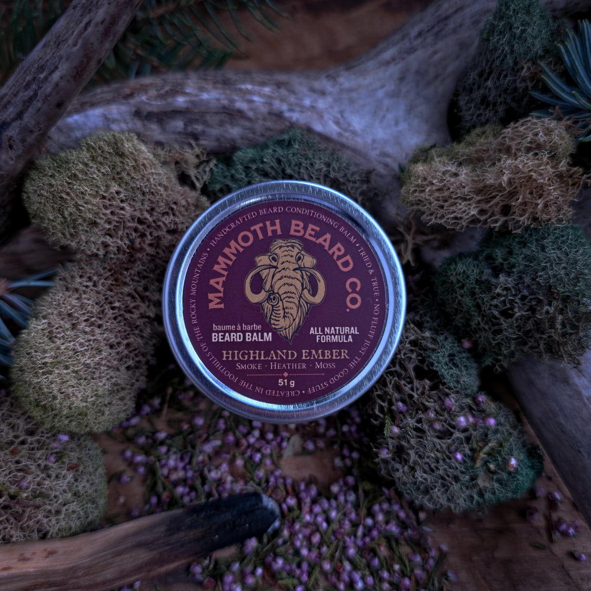 Close-up, top-down view of Mammoth Beard Co. Highland Ember beard balm tin nestled among green moss, pine, and heather on a wooden backdrop.