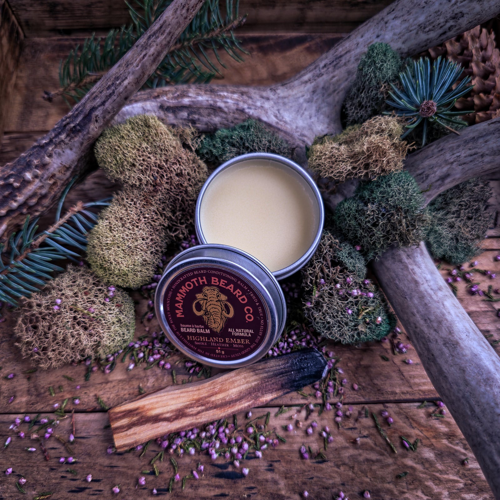 Open tin of Mammoth Beard Co. Highland Ember beard balm on rustic wood, styled with moss, pine sprigs, antler, and a charred wood stick.