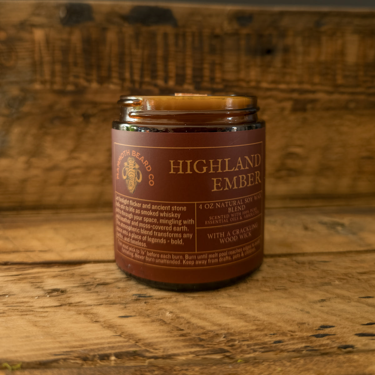 Highland Ember soy wax candle in amber jar with wood wick, set against a rustic wooden background engraved with the Mammoth Beard Co. logo.