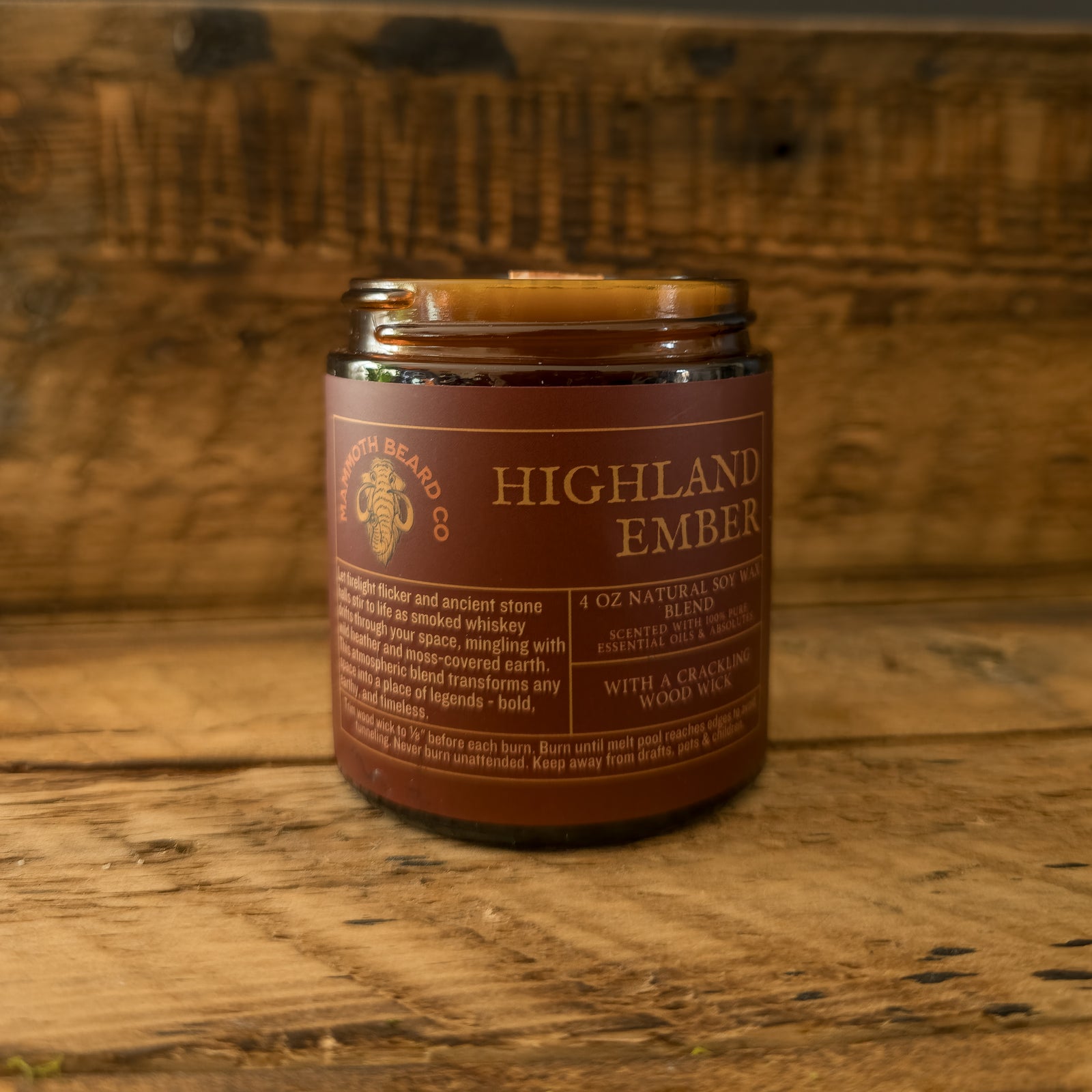 Highland Ember soy wax candle in amber jar with wood wick, set against a rustic wooden background engraved with the Mammoth Beard Co. logo.