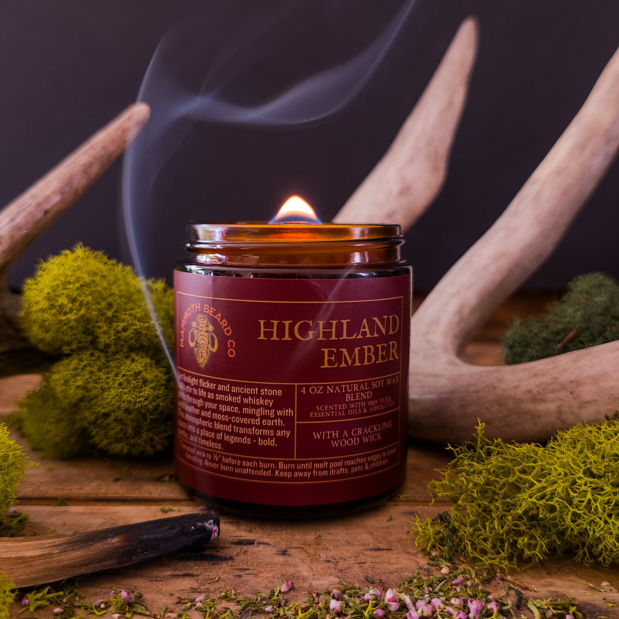 Highland Ember candle by Mammoth Beard Co. burning with a crackling wood wick, surrounded by moss and antlers, evoking a rustic smoky whiskey and moss-covered earth scent.