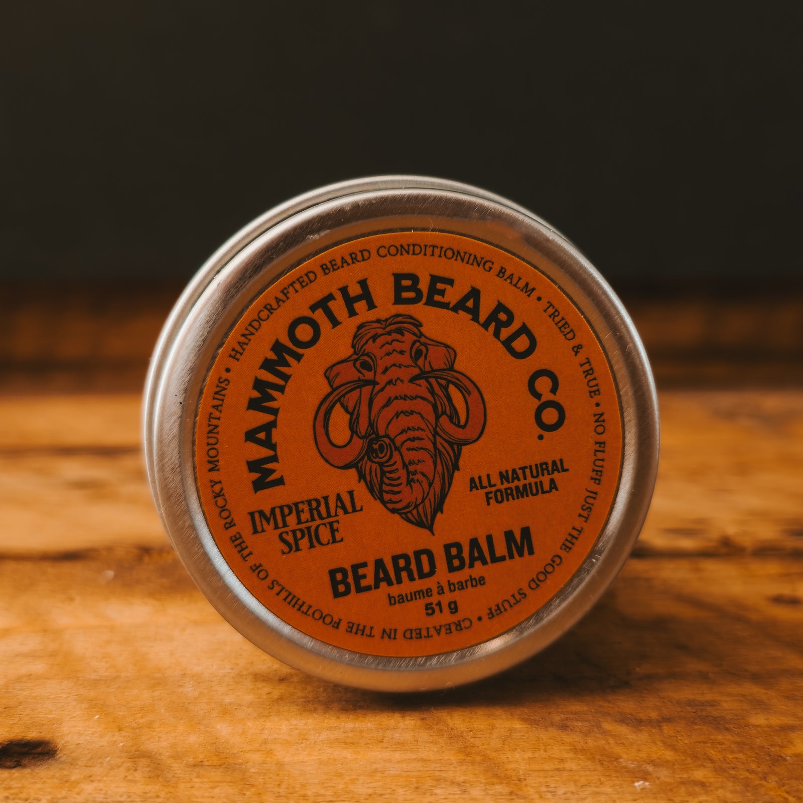 A round metal tin of Mammoth Beard Co. Imperial Spice Beard Balm, featuring a prominent orange label, resting on a wooden surface.