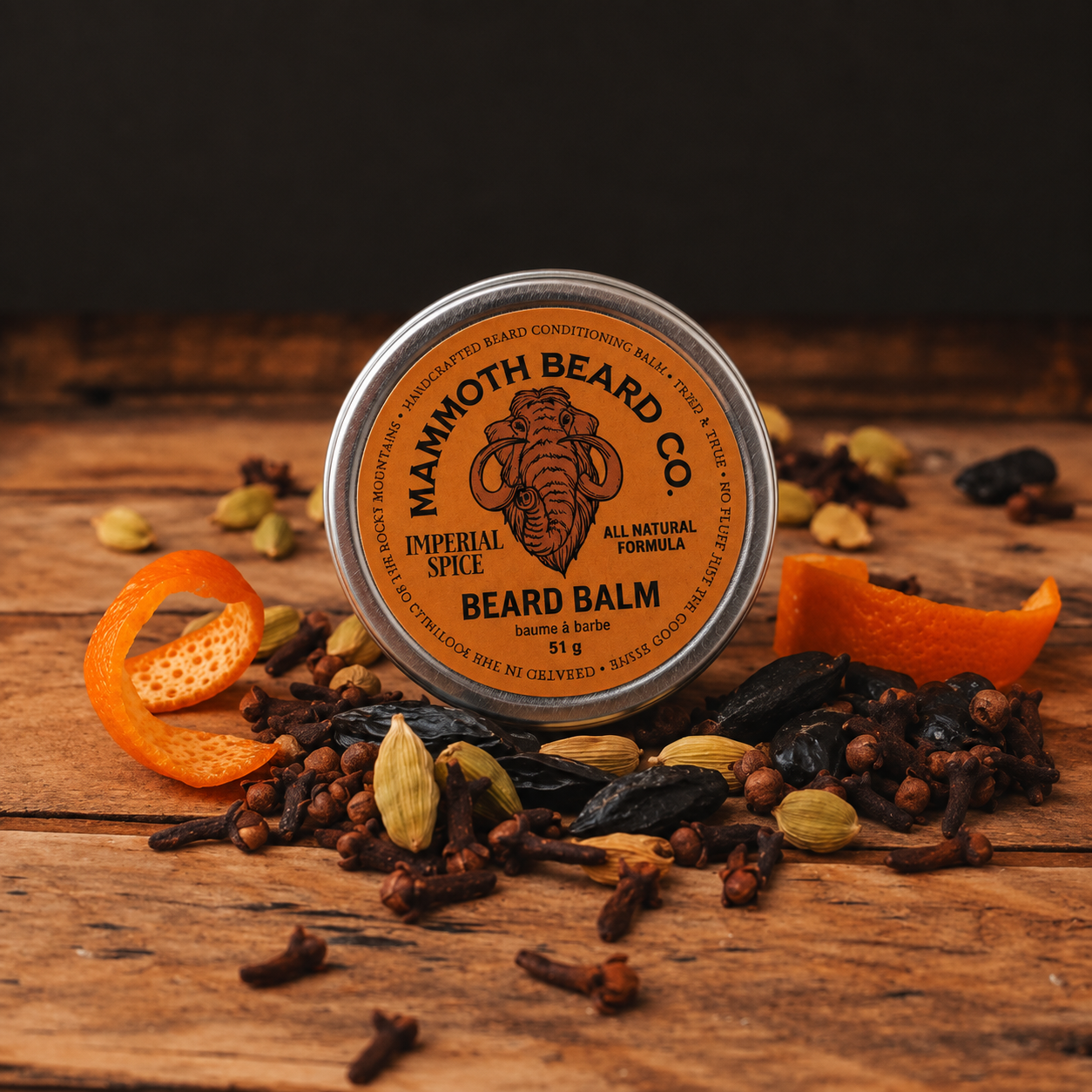 A round tin of Mammoth Beard Co. Imperial Spice Beard Balm sits on a rustic wooden surface surrounded by tonka beans, cardamom pods, cloves, and orange peel slices against a dark background.