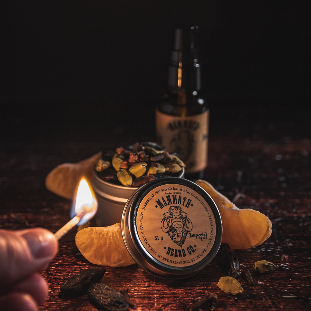 Imperial Spice Beard Balm tin styled with spices and citrus peel, with Mammoth Beard Co. beard oil in the background and a lit match in the foreground for a moody, warm atmosphere.
