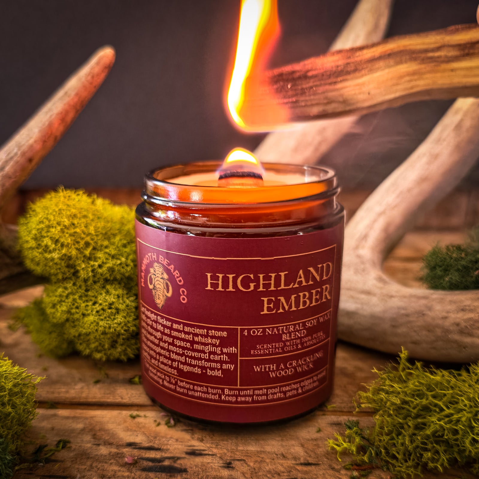 Close-up of the Highland Ember soy wax candle being lit with a match, featuring a crackling wood wick and deep earthy design with moss and antlers in the background.
