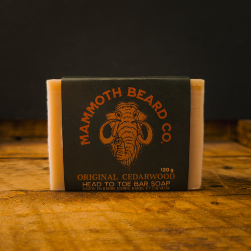 A bar of natural soap wrapped in a dark green label with orange text reading 'Mammoth Beard Co. Original Cedarwood' and a mammoth illustration, standing on a rustic wooden surface against a dark background.