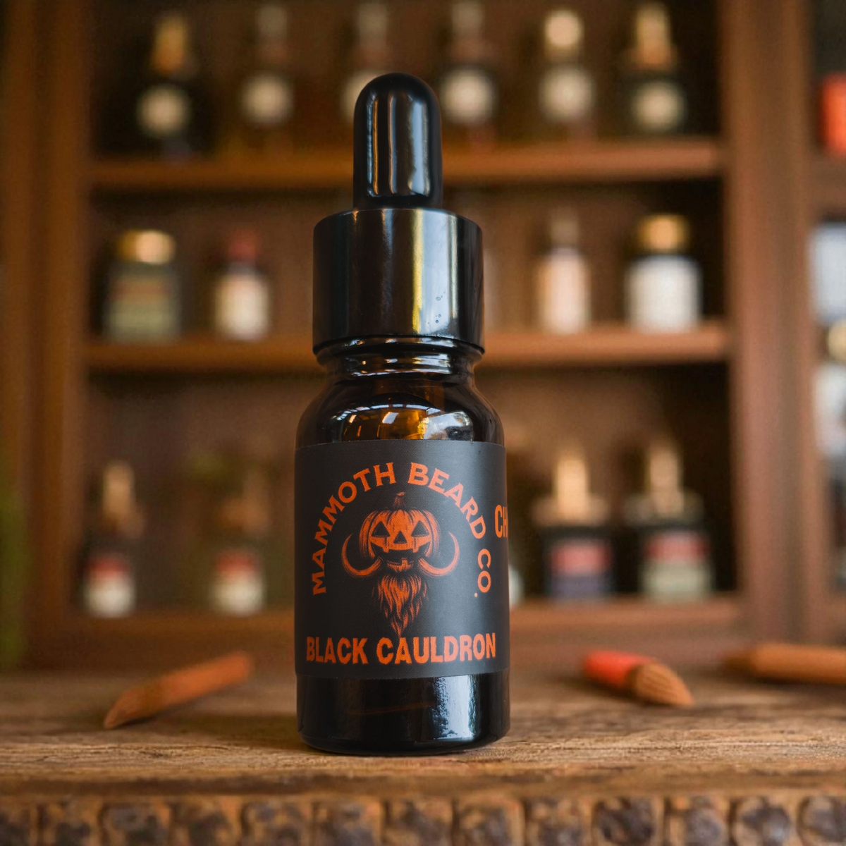 10ml bottle of Mammoth Beard Co. Black Cauldron beard oil displayed on a rustic wooden surface with a blurred background of an apothecary shelf. Halloween themed label featuring a mammoth-pumpkin logo in orange and black.