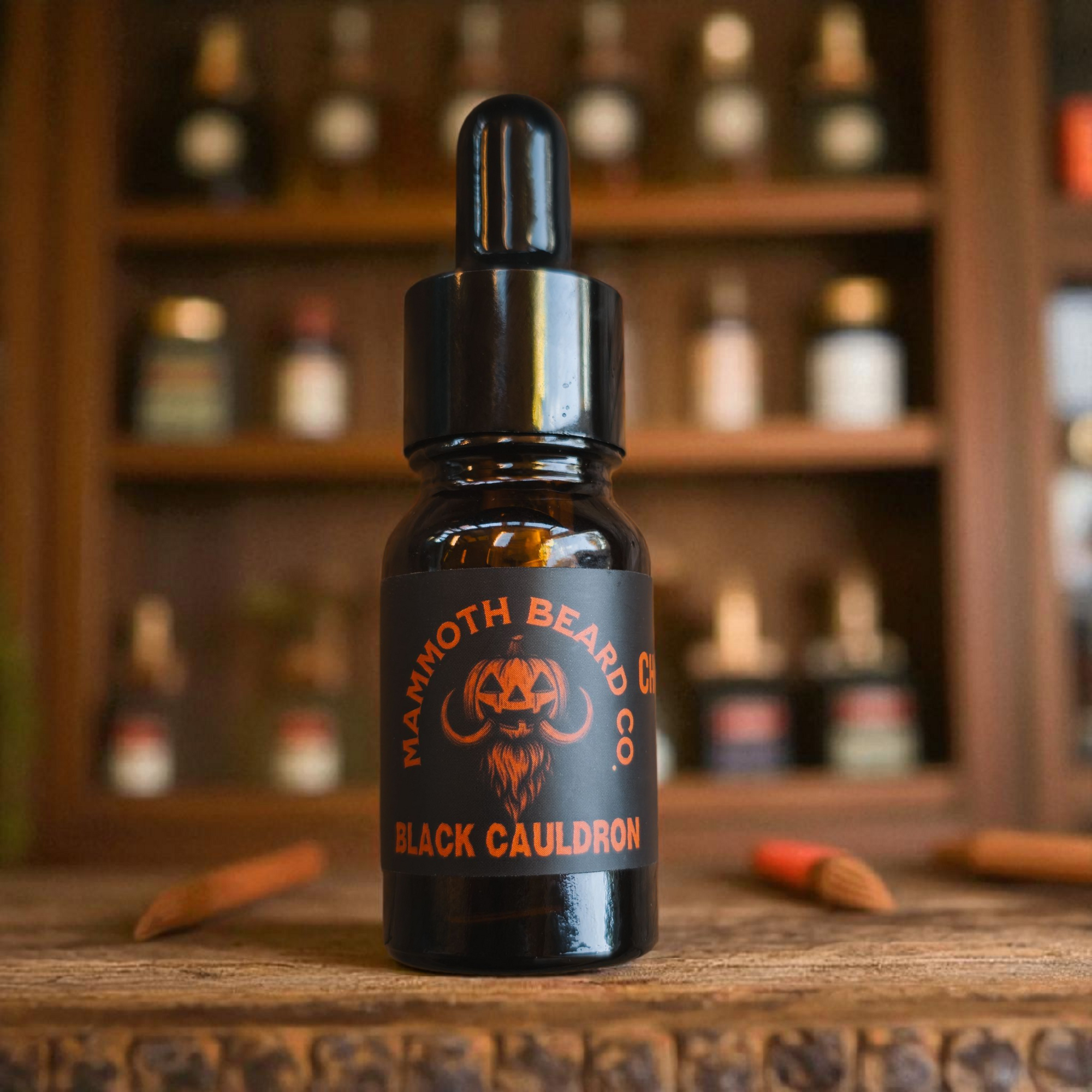 10ml bottle of Mammoth Beard Co. Black Cauldron beard oil displayed on a rustic wooden surface with a blurred background of an apothecary shelf. Halloween themed label featuring a mammoth-pumpkin logo in orange and black.
