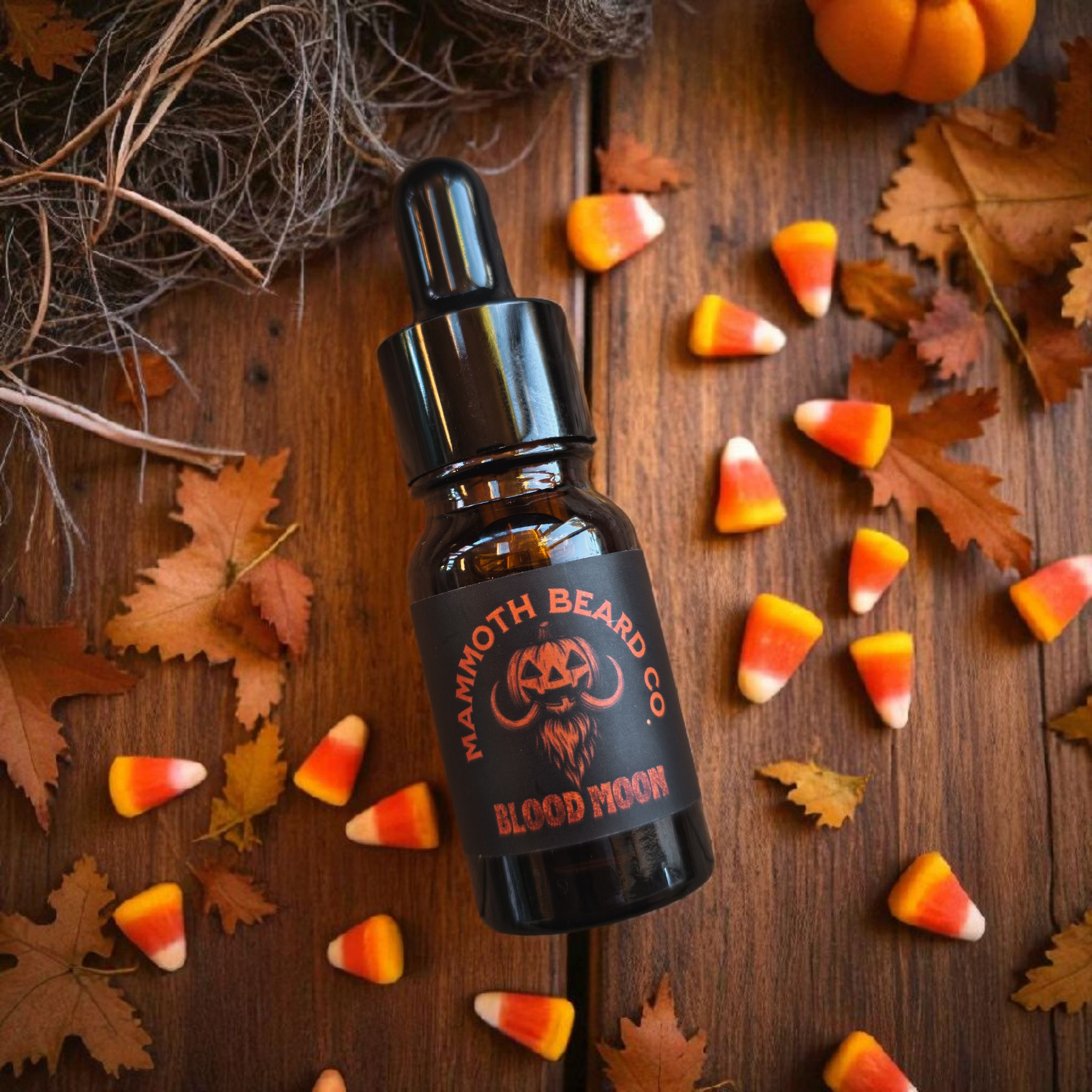 10ml bottle of Mammpth Beard Co. Blood Moon beard oil lying on  wooden surface surrounded by autumn leaves, candy corn, and a small pumpkin. Halloween-themed label with orange and black pumpkin-mammoth beard logo.