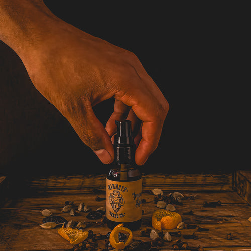 Hand reaching for a bottle of Mammoth Beard Co. beard oil on a rustic wooden surface surrounded by clove, cardamom, and dried orange peel, highlighting the natural, nutrient-rich Hazelnut & Hemp formula.