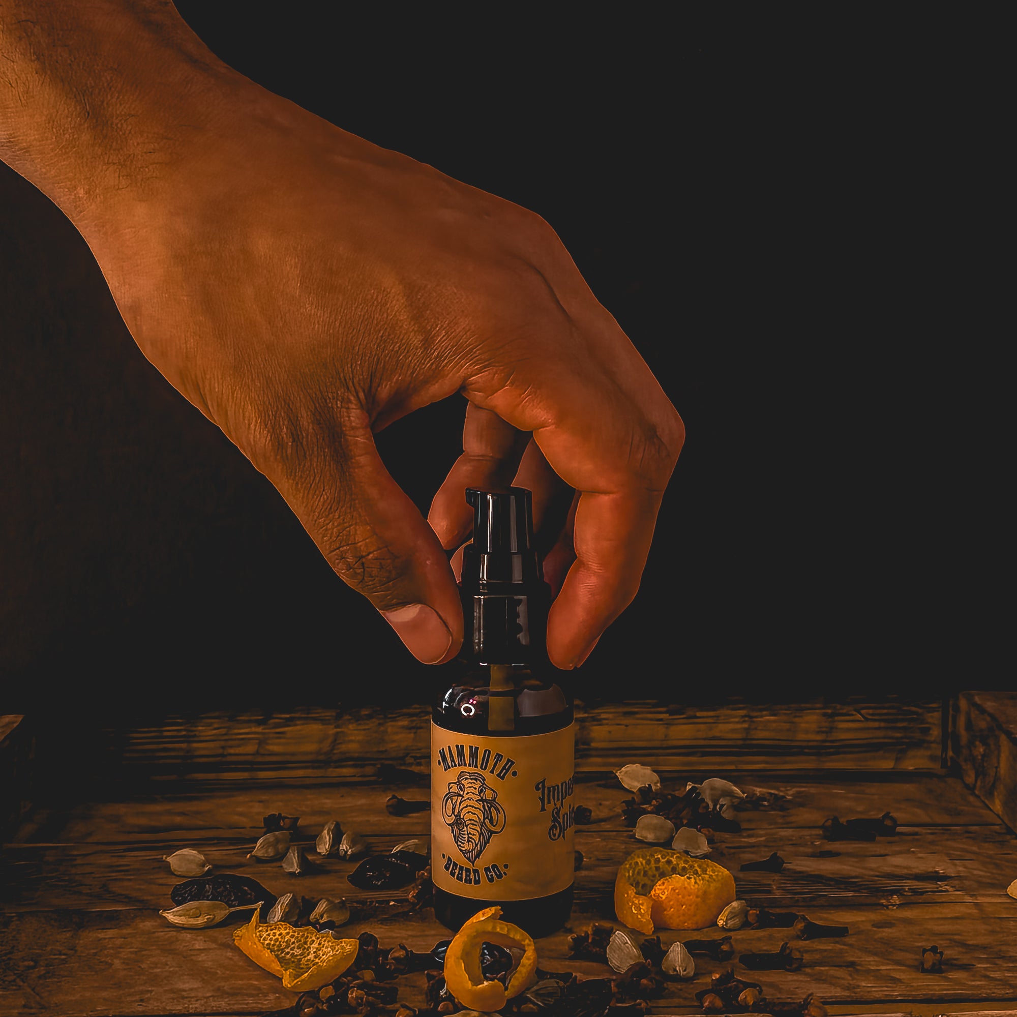Hand reaching for a bottle of Mammoth Beard Co. beard oil on a rustic wooden surface surrounded by clove, cardamom, and dried orange peel, highlighting the natural, nutrient-rich Hazelnut & Hemp formula.