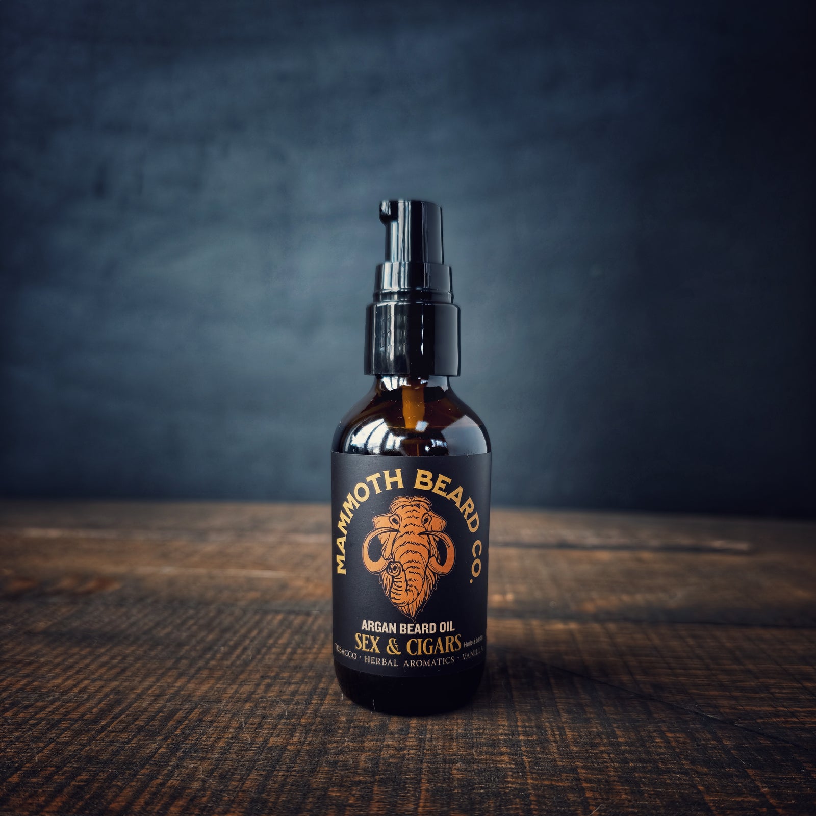 A bottle of Mammoth Beard Co. Sex & Cigars Argan Beard Oil on a rustic wood surface with a dark background, highlighting the premium amber glass and mammoth logo.