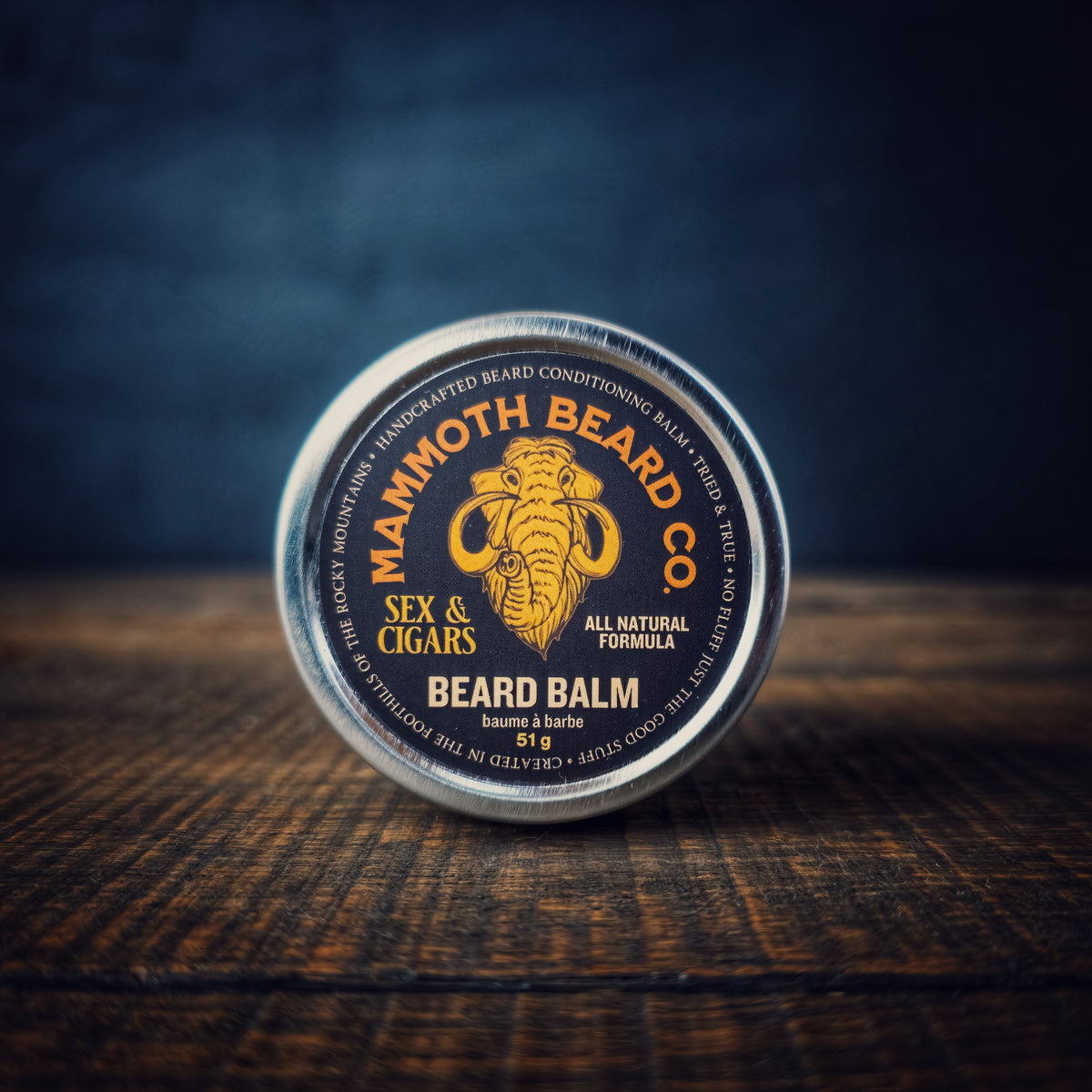 A tin of Mammoth Beard Co. Sex &amp; Cigars Beard Balm photographed on a wood tabletop with a dark blurred background, emphasizing the clean black and gold design.
