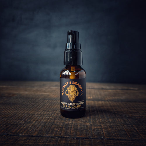 Mammoth Beard Co. Sex & Cigars Hazelnut & Hemp Beard Oil displayed on a wooden table with a dark moody backdrop, showing the amber bottle and bold black label.