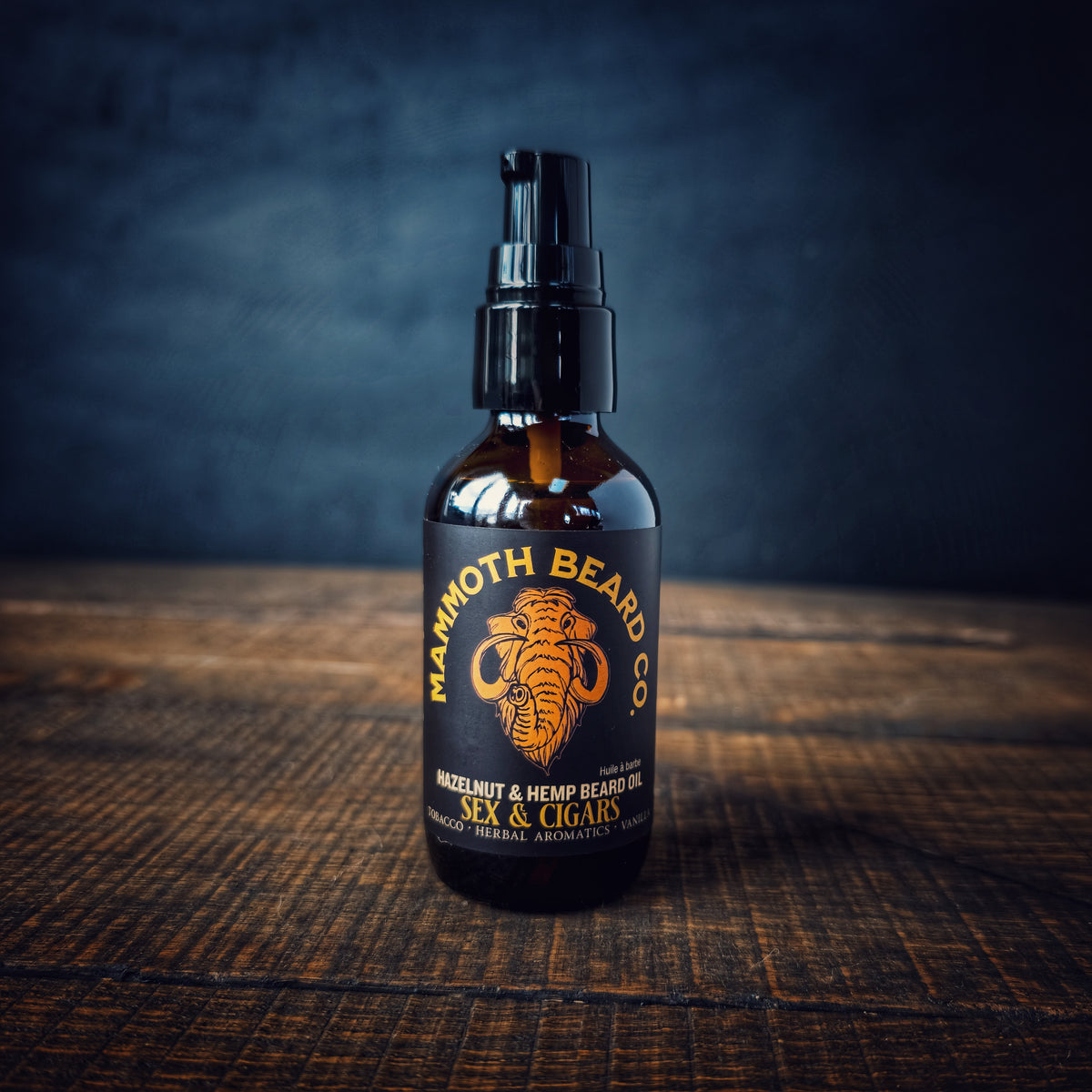 Mammoth Beard Co. Sex &amp; Cigars Hazelnut &amp; Hemp Beard Oil displayed on a wooden table with a dark moody backdrop, showing the amber bottle and bold black label.