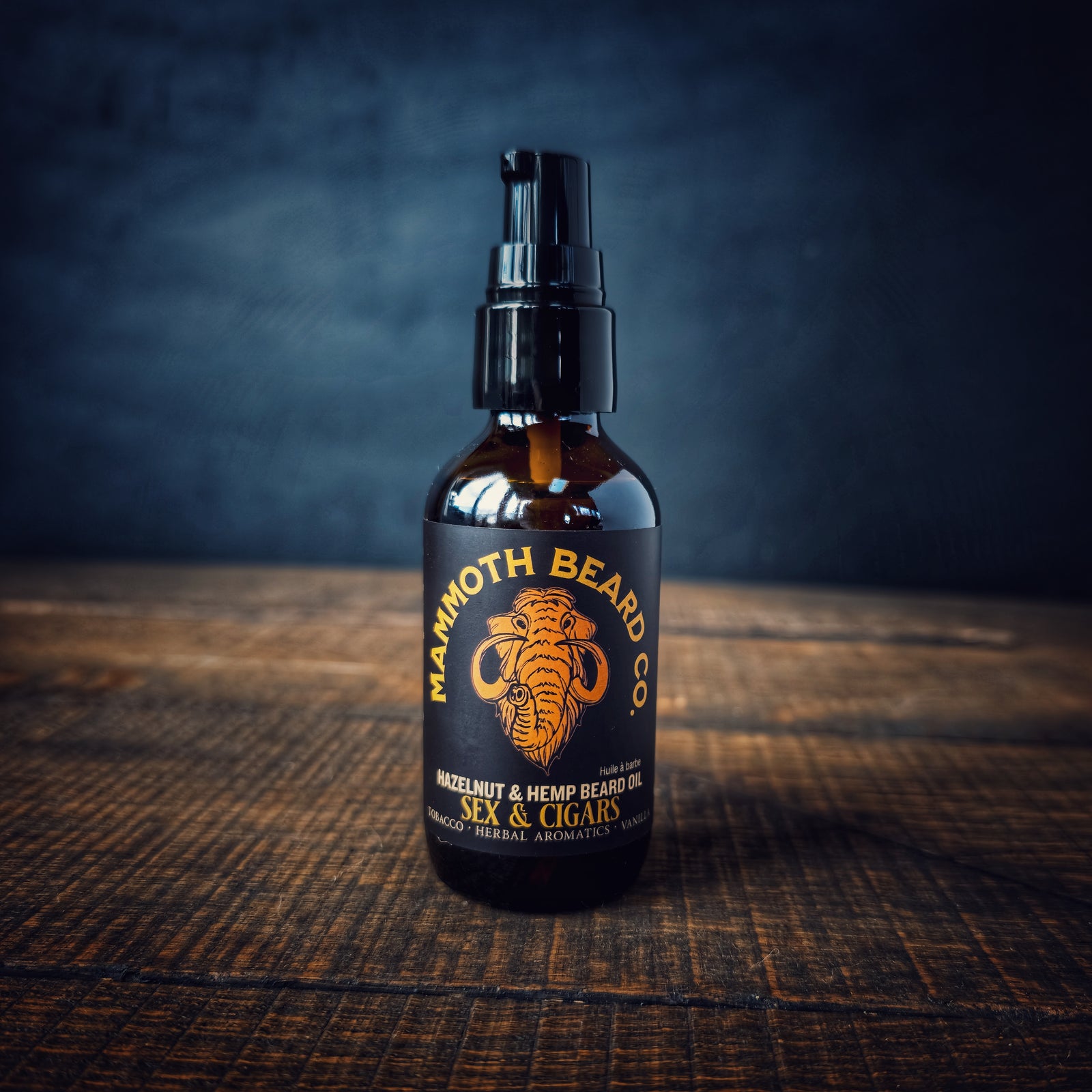 Mammoth Beard Co. Sex & Cigars Hazelnut & Hemp Beard Oil displayed on a wooden table with a dark moody backdrop, showing the amber bottle and bold black label.