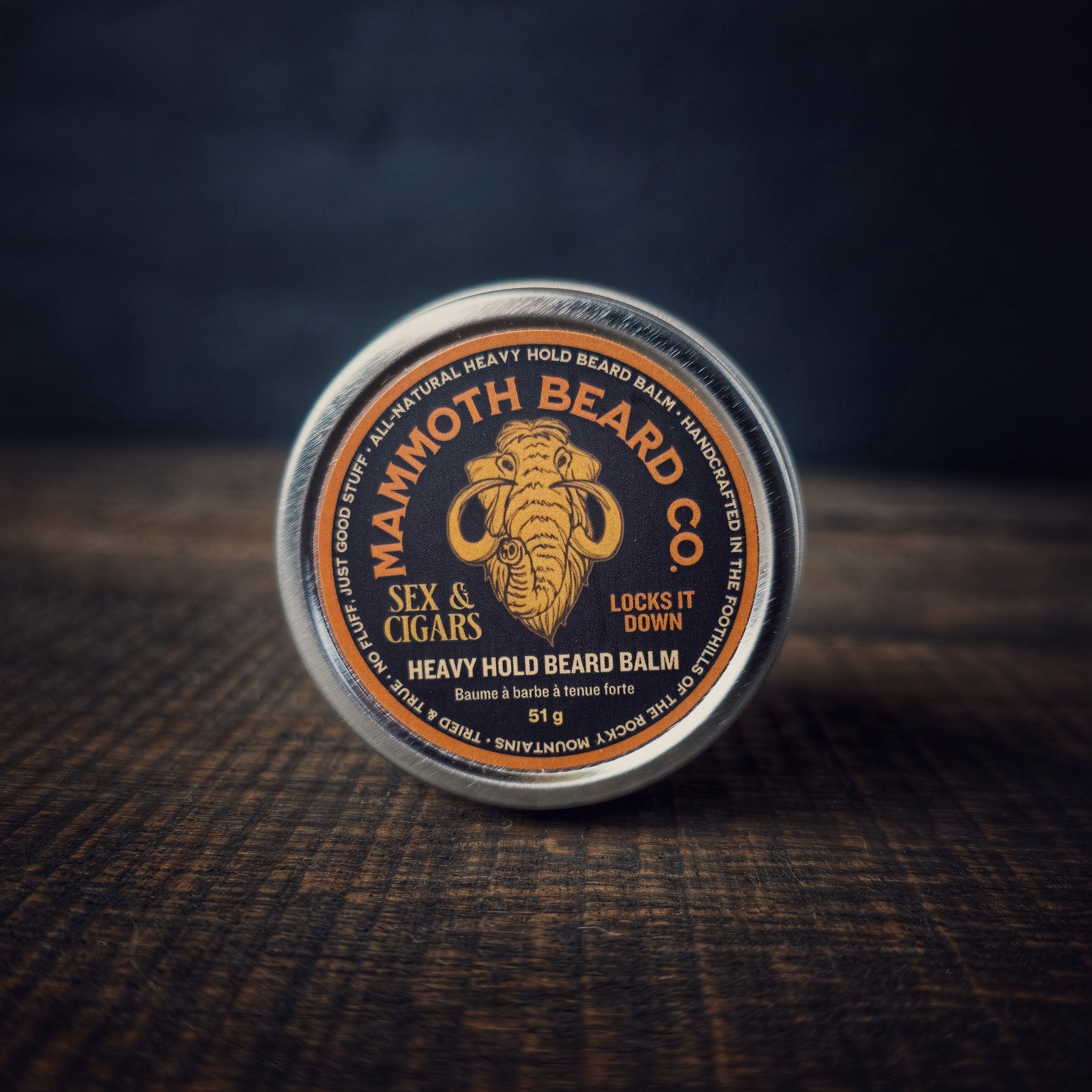 Mammoth Beard Co. Sex & Cigars Heavy Hold Beard Balm tin resting on a wooden surface, featuring the mammoth logo and “Locks It Down” text on the label.