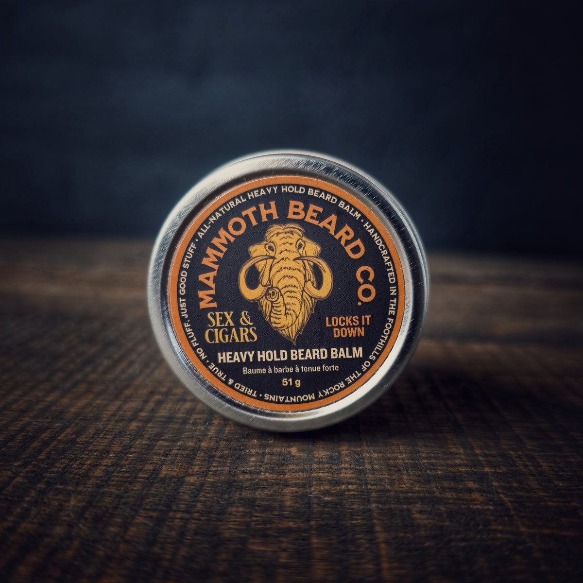 Mammoth Beard Co. Sex &amp; Cigars Heavy Hold Beard Balm tin resting on a wooden surface, featuring the mammoth logo and “Locks It Down” text on the label.