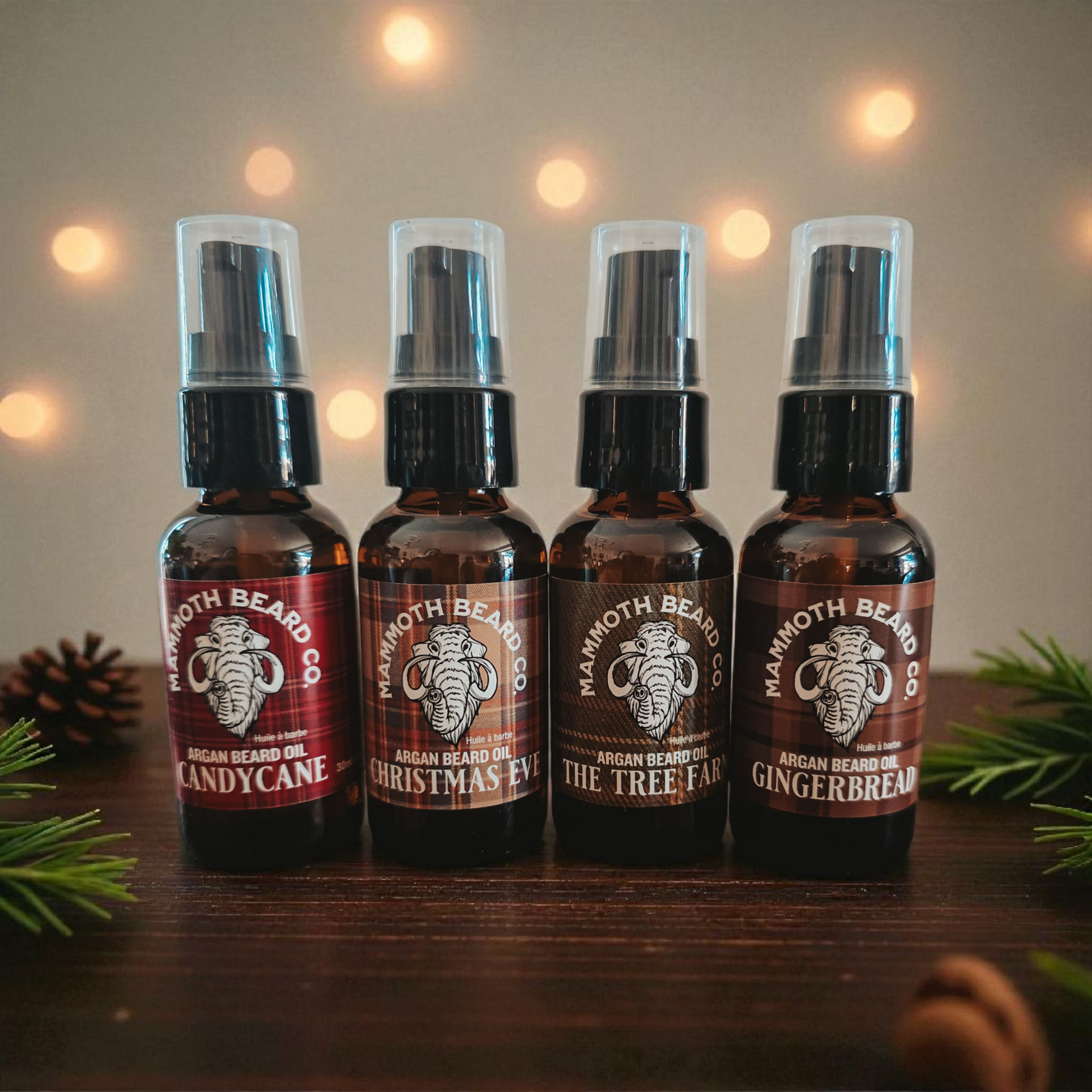Four Mammoth Beard Co Christmas Argan Beard Oils—Candy Cane, Christmas Eve, The Tree Farm, and Gingerbread—arranged on a wooden surface with festive lights in the background.