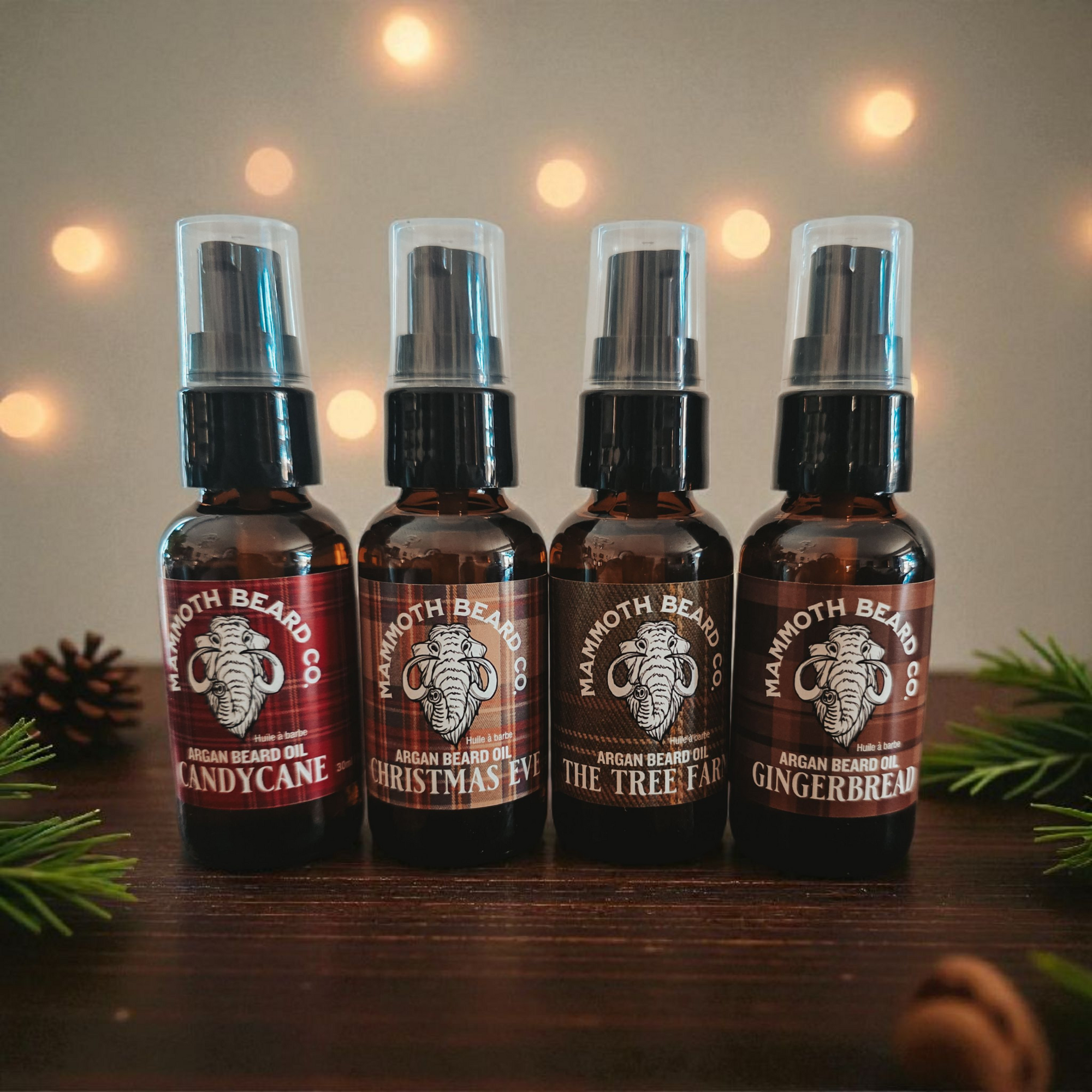 Four Mammoth Beard Co Christmas Argan Beard Oils—Candy Cane, Christmas Eve, The Tree Farm, and Gingerbread—arranged on a wooden surface with festive lights in the background.