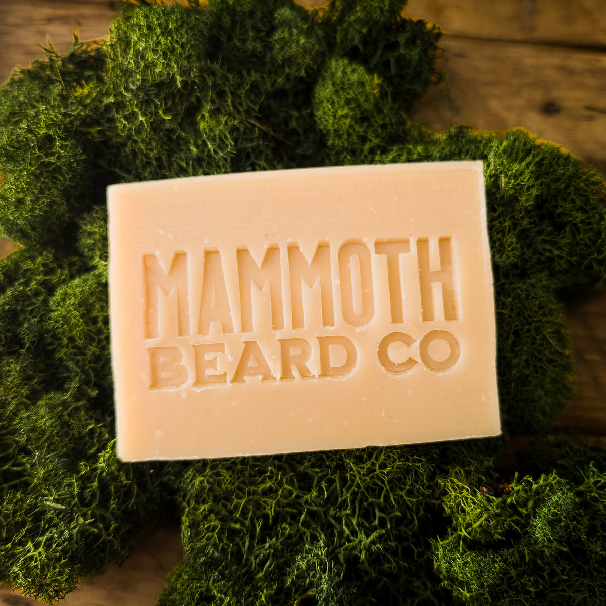 A rectangular bar of pale beige soap embossed with 'MAMMOTH BEARD CO' resting on a bed of green moss over a rustic wooden surface.