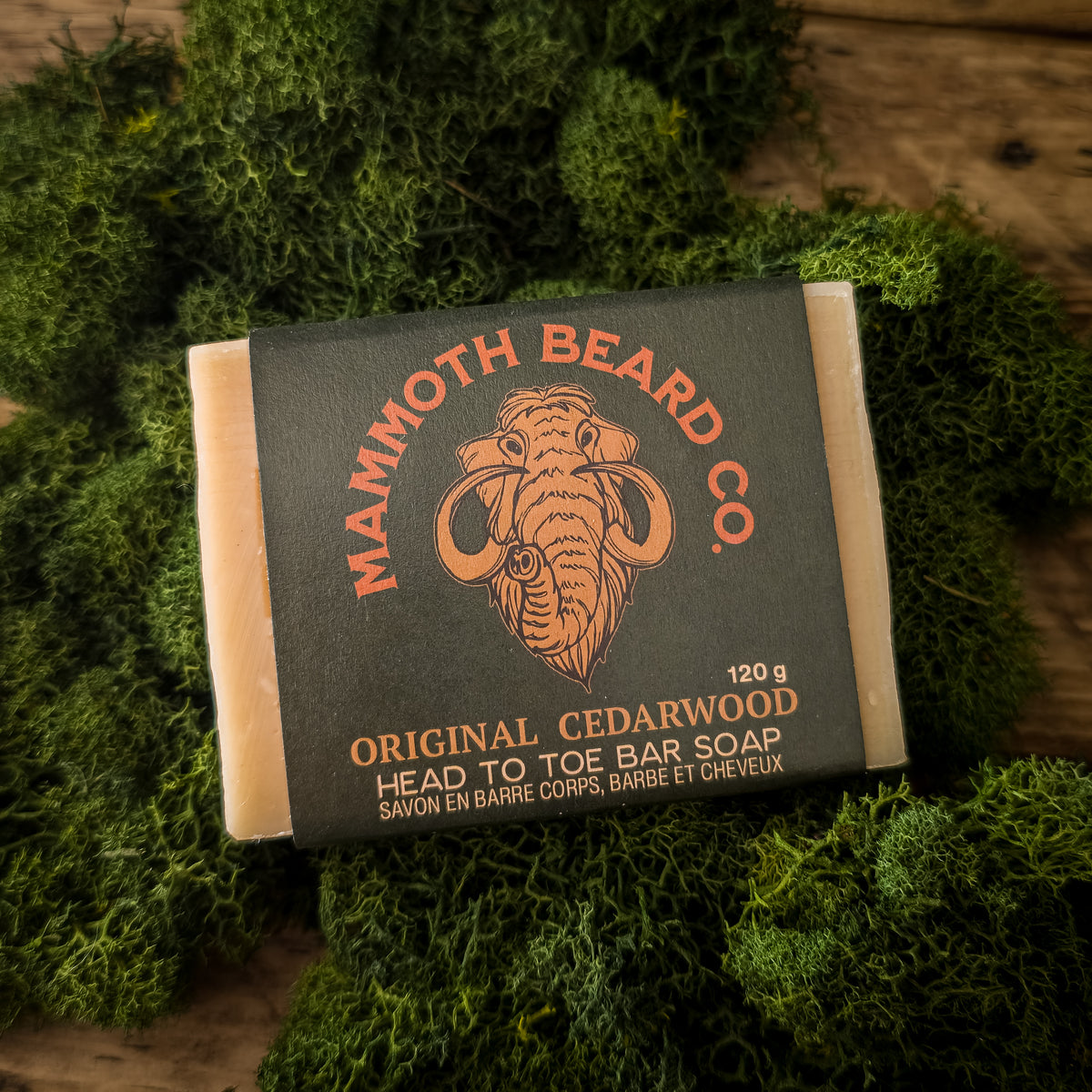 A bar of Mammoth Beard Co. Original Cedarwood Head to Toe Bar Soap resting on a bed of green moss, with a rustic wooden surface visible in the background.