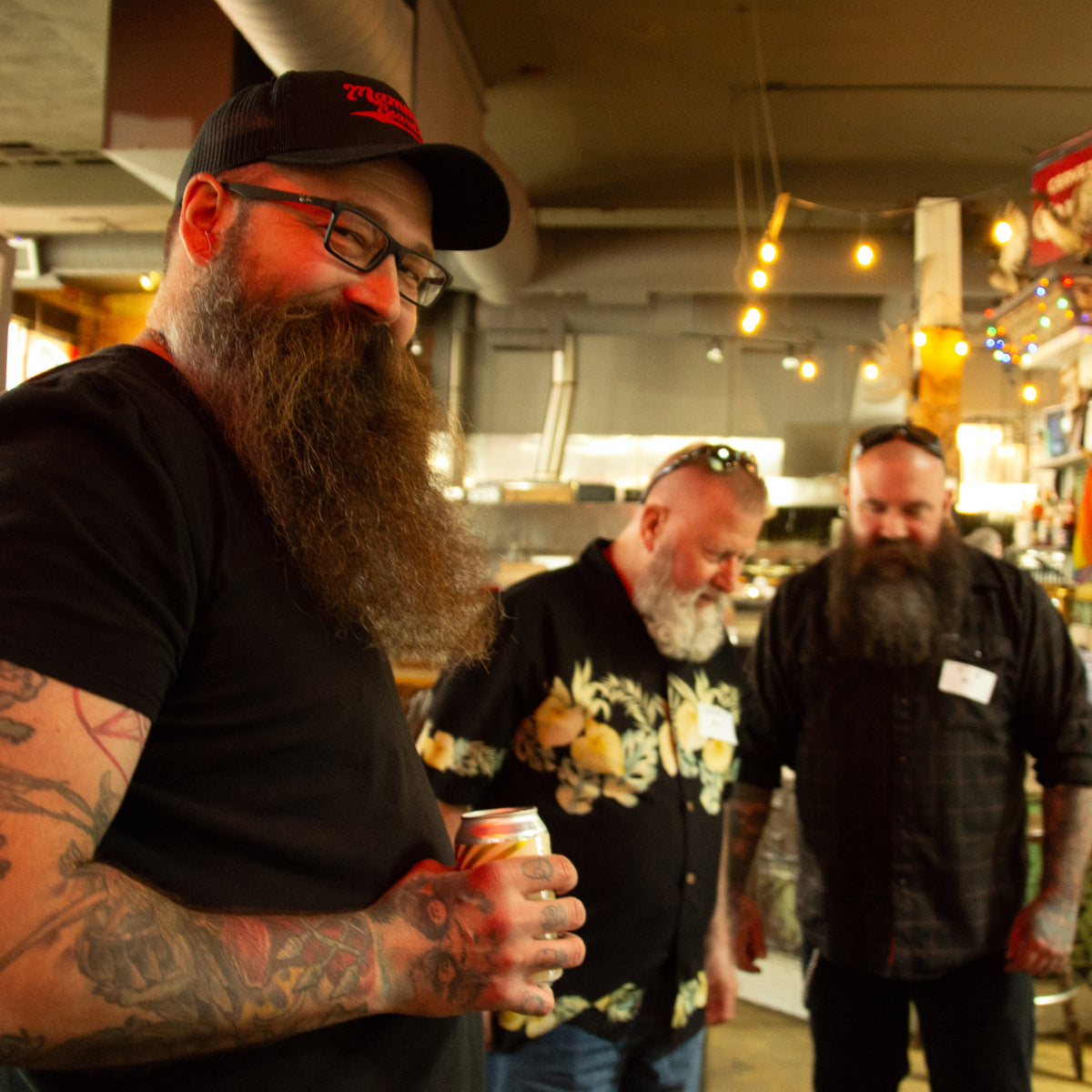 Three bearded men standing in a dimly lit indoor venue or bar. The man in the foreground on the left wears a black t-shirt, black cap, and glasses while holding a beverage can.