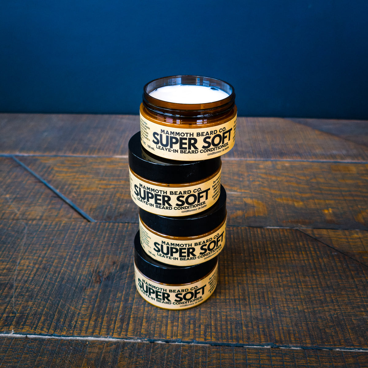 Four stacked amber jars of Mammoth Beards Co. Super Soft Beard Conditioner on a dark rustic wooden table against a dark blue background.